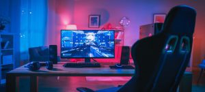 Powerful Personal Computer Gamer Rig with First-Person Shooter Game on Screen. Monitor Stands on the Table at Home. Cozy Room with Modern Design is Lit with Pink Neon Light.