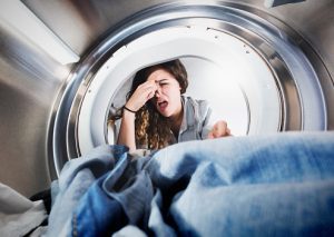 A lovely young woman grimaces, holding her nose as she reaches into a tumble dryer to remove some smelly laundry. Seen from inside the drier.