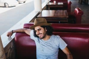 Adult casual man in hat sitting on sofa in outside cafe on terrace looking away and relaxing