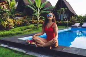 Joyful long haired lady in sunglasses with flower in hairs smiling and looking at camera while relaxing on poolside in exotic yard